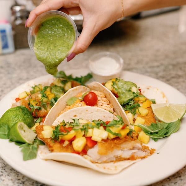 A hand drizzles green sauce over three Remedy Diner fish tacos with mango salsa and lime.