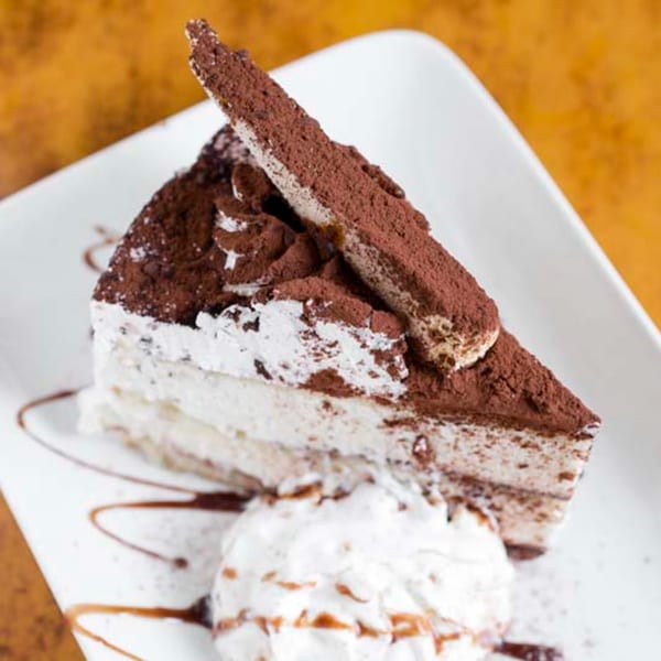 A slice of tiramisu with whipped cream and chocolate drizzle on a white plate.