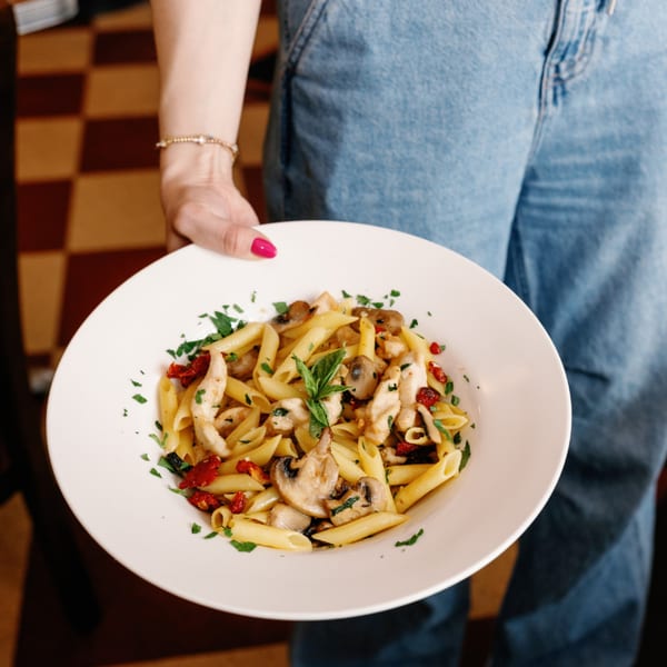 A person in blue jeans holds a white plate of penne pasta with chicken, mushrooms, and sun-dried tomatoes.