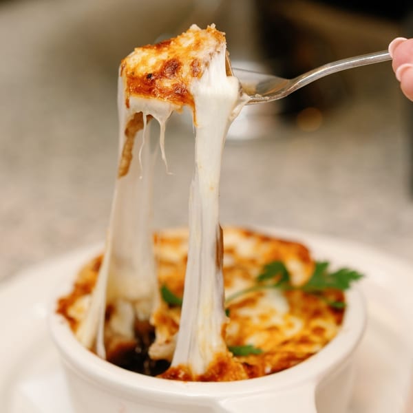 A fork lifts a piece of cheesy French onion soup from a white bowl, stretching the melted cheese.