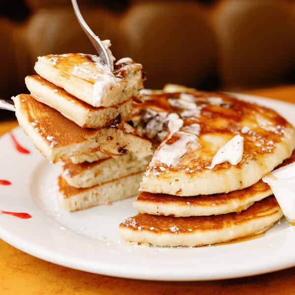 A fork lifts a piece of fluffy Remedy Diner pancake, topped with syrup, butter, and powdered sugar.