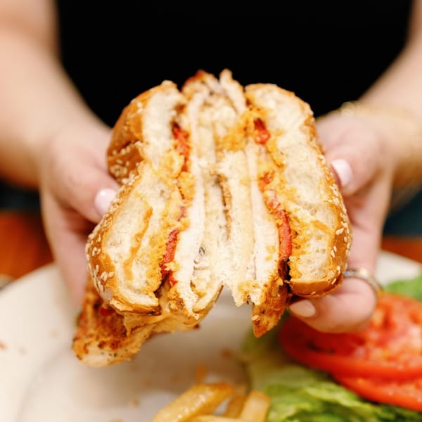Hands holding a Remedy Diner chicken sandwich, cut in half, revealing layers of chicken and red peppers.