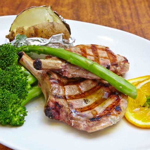 Grilled lamb chops with asparagus, broccoli, a baked potato, and orange slices on a white plate.