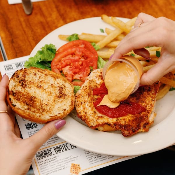 A diner customer squeezes sauce onto a Remedy Diner chicken sandwich with fries and salad.