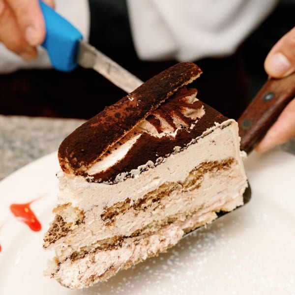 A hand serving a slice of tiramisu cake with cocoa powder and ladyfingers on a white plate.