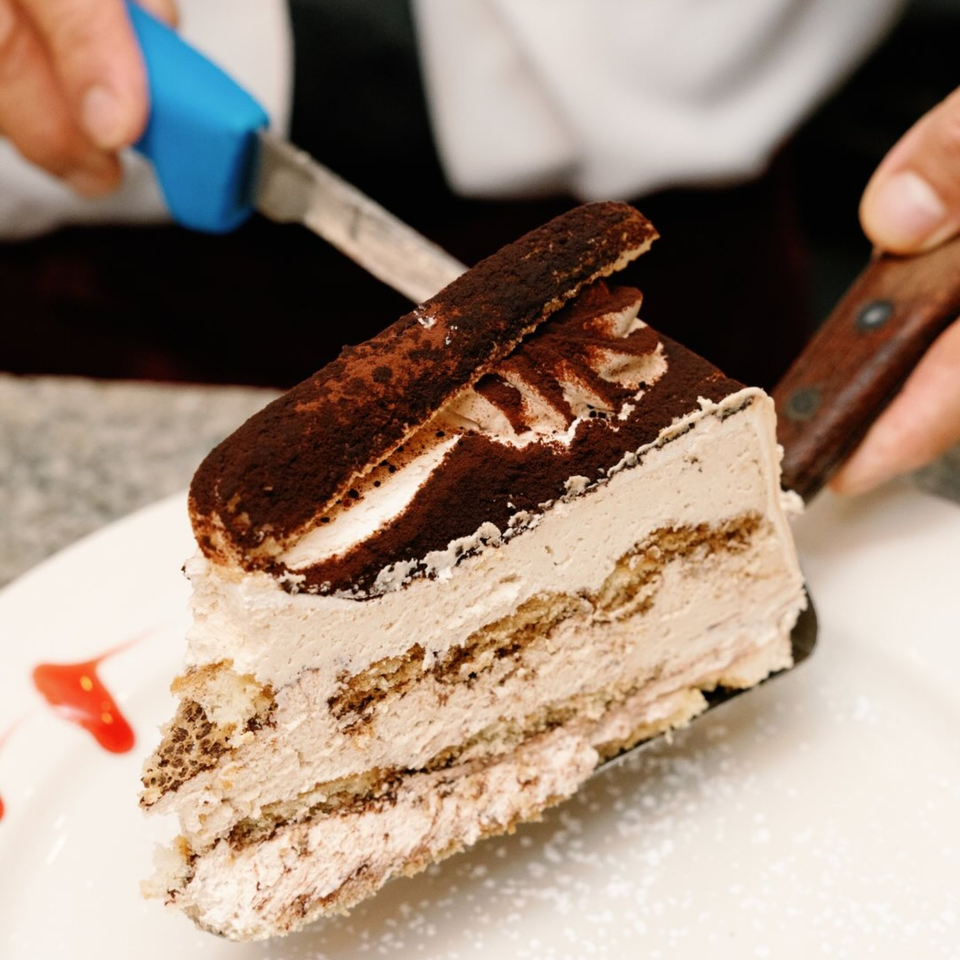 A hand serving a slice of tiramisu cake with cocoa powder and ladyfingers on a white plate.