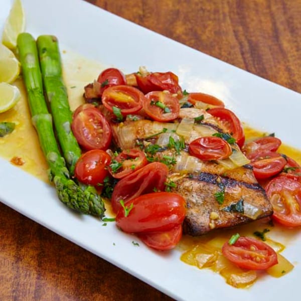 Grilled fish topped with cherry tomatoes and onions, served with asparagus and lemon wedges.