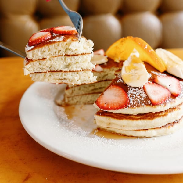 A fork lifts a pancake from a stack of Remedy Diner pancakes with strawberries, bananas, and syrup.