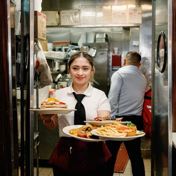 A smiling Remedy Diner waitress carries plates of pancakes, quesadillas, and fries from the kitchen.