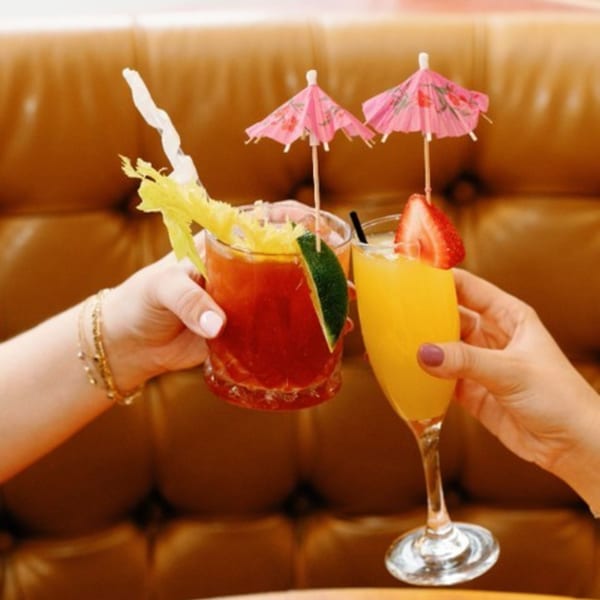 Two hands clinking glasses of a Bloody Mary and a Mimosa, garnished with umbrellas, at Remedy Diner.