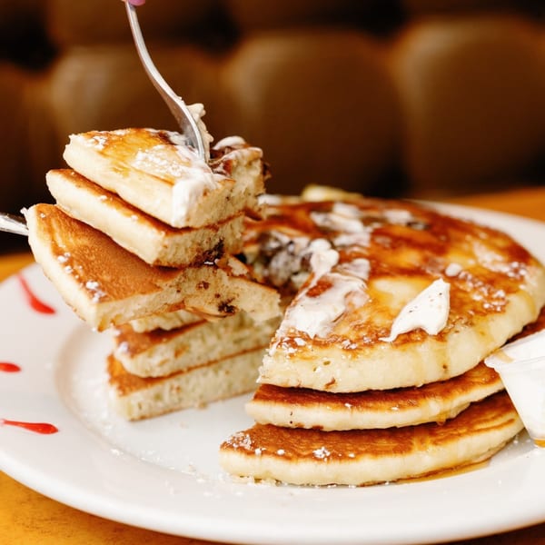 A fork lifts a piece of fluffy Remedy Diner pancake, drizzled with syrup and powdered sugar.