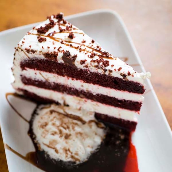 A slice of red velvet cake with white frosting and chocolate drizzle on a white plate.