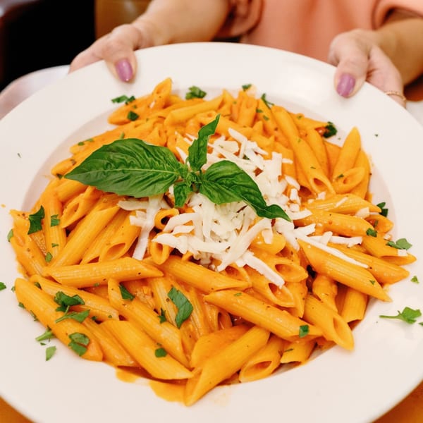 Hands holding a white plate of penne pasta with red sauce, shredded cheese, and basil.
