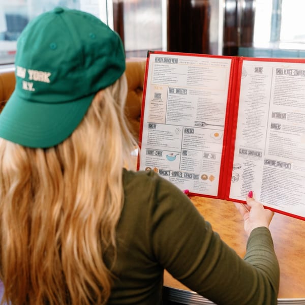 A person in a green hat reads a Remedy Diner menu, featuring brunch items like omelets and pancakes.