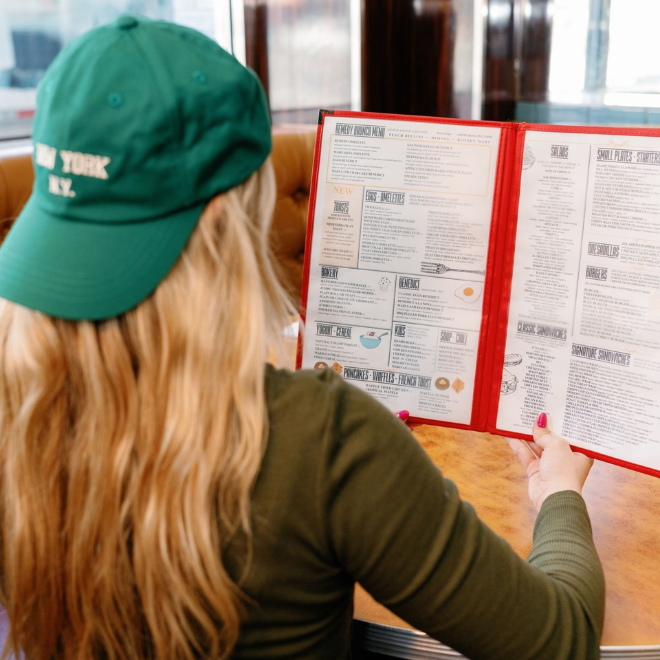 A person in a green hat reads a Remedy Diner menu, featuring brunch items like omelets and pancakes.